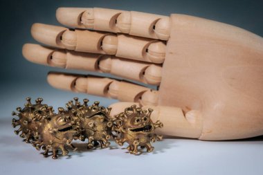 Tahta bir mankenin eli birkaç bronz virüs şekilli heykelciğin üzerinde asılı. Yüzleri aydınlık bir yüzeyde..