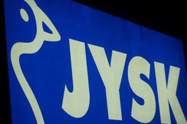 Parlak, mavi-beyaz bir JYSK logosu, gece çekilmiş, bir perakende mağazasının vitrininde tanınabilir stilize edilmiş bir kaz..