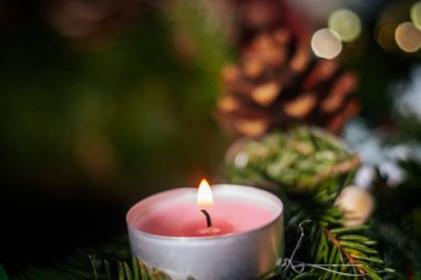 Parlayan turuncu bir mum yeşil çam iğnelerinin üzerinde nazikçe yanarak sıcak, şenlikli ve samimi bir tatil atmosferi yaratır. Kopyalama alanı olan koyu arkaplan.