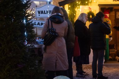 Bir grup sıcak giyimli insan, açık hava Noel standında, ışık, yeşillik ve tatil tabelasıyla sırada bekliyor..