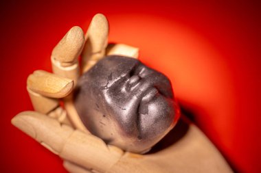 Parlak, yüz şeklinde bir vudu bebeği kafası, parlak kırmızı arka planda dramatik bir ışık altında bir insan eli tarafından tutuluyor. Parlak dokuyu ve yontulmuş yüz özelliklerini vurgulamak.