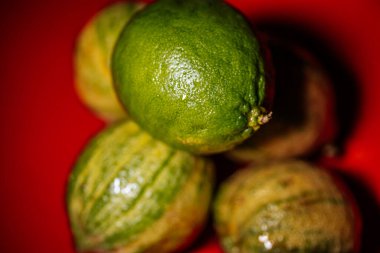 Parlak, kırmızı bir zemin üzerinde kabuklu yeşil ve sarı kaffir limonlu makro yakın çekim. Olgunluk farklılıklarını ve doğal yüzey detaylarını vurguluyor..