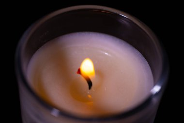 Açık cam bir kavanozda, yumuşak turuncu bir alev ve koyu bir arka planda sıcak balmumu olan bir mumun yakın çekimi..