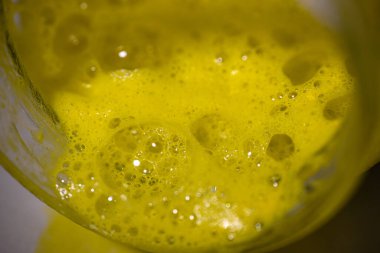 Cam bir kavanozda ince kabarcıkları olan yoğun sarı köpük ince yansımaları olan parlak bir doku yaratıyor ve arka planda çok az yer alıyor..