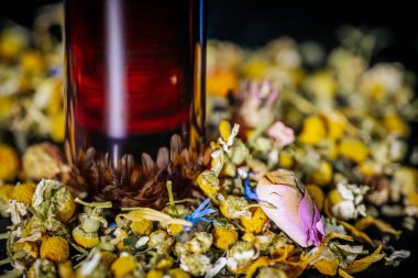 Kahverengi şapkalı dikey kırmızı şişe koyu arkaplanda kurumuş papatya, gül tomurcukları ve bitkisel yaprakların karışımı duruyor..