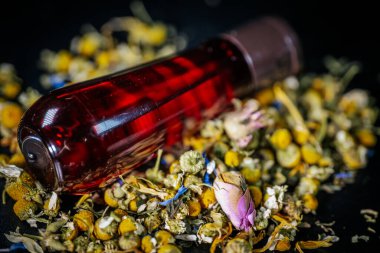 Kahverengi şapkalı dikey kırmızı şişe koyu arkaplanda kurumuş papatya, gül tomurcukları ve bitkisel yaprakların karışımı duruyor..