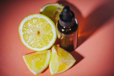 Kahverengi cam bir şişe ve damlatan yumuşak pembe bir yüzeyde taze, sulu limon dilimlerinin yanında oturur. Kompozisyonda doğal sağlık, cilt bakımı veya sağlıkla ilgili konular vurgulanmaktadır..