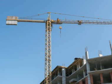 Bir inşaat vinci, açık mavi gökyüzüne karşı inşa halinde olan monolitik bir iskeletin yanında duruyor.