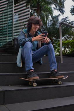 Genç bir adam merdivenlerde oturmuş akıllı telefonuna bakıyor. Ayaklarının dibinde bir kaykay var.