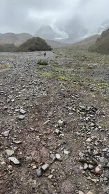 Laguna Humantay 'a çıkan kayalık arazide sisli bir dağ yolu esiyor. Uzaktaki yürüyüşçüler bulutların ötesindeki turkuaz lagüne doğru ilerliyorlar, yüksek tepelerle çevrili. Yüksek kalite 4k görüntü.