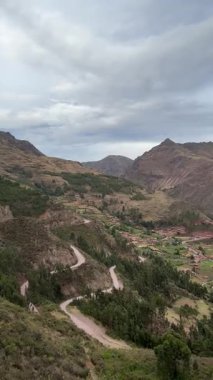 Bulutlu gökyüzünün altındaki yeşil dağ vadisindeki küçük bir köye giden dolambaçlı bir zigzag yolunun panoramik manzarası. Kırsal arazileri ve tarım alanlarını ön plana çıkarıyor. Yüksek kalite 4k görüntü