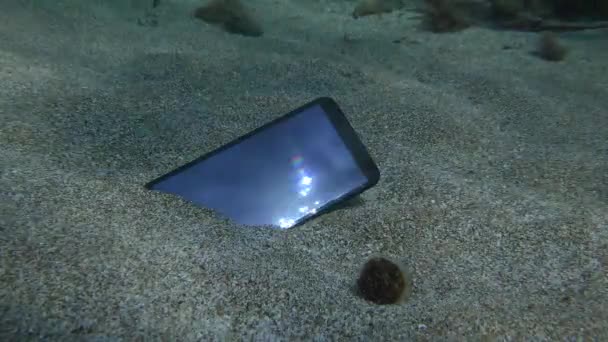 I raggi del sole riflessi dallo schermo di uno smartphone smarrito sdraiato su un fondo sabbioso, colpo subacqueo. — Video Stock