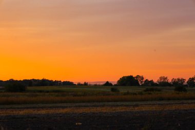Nefes kesen bir şafak ya da gün batımı manzarası ön planda, koyu ve altın renklerin hakim olduğu kırsal bir arazide sergilenir.. 