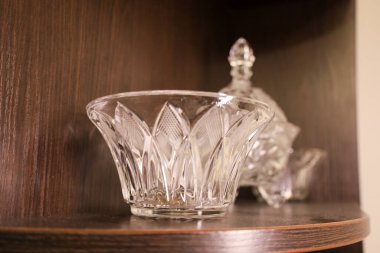 A crystal decorative vase-bowl with elegant engraving stands on a dark wooden shelf, creating a refined texture contrast. The glittering glassware with sharp facets elegantly highlights the home interior.