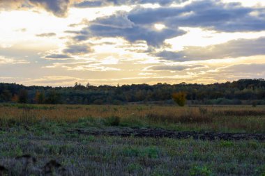 A sweeping rural landscape showcases a dynamic sky with layered grey and white clouds, through which sunlight breaks, creating a bright glow.