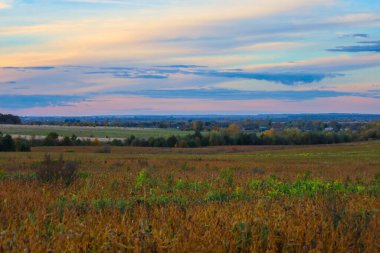 A vast rural landscape at twilight showcases a wide sky with soft hues of blue, pink, and orange over rolling fields. The foreground is dominated by dry, golden-brown vegetation, adding an autumnal feel.
