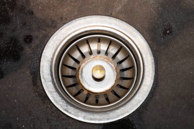 Bu detaylı yakın plan, karanlık ve ıslak bir yüzeyle çevrili bir lavaboda, endüstriyel tasarımı, kullanım ve zaman işaretlerini vurgulayan bir metal drenaj açılışını gösteriyor..