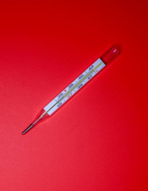 Açık bir şekilde görülebilen bir sütuna sahip cıvalı bir tıbbi termometre parlak kırmızı bir arka planda çapraz olarak yatar. Ölçüm aracı hastalığı, yüksek sıcaklığı ve tıbbi kontrol ihtiyacını simgeliyor..
