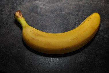 Hafif kıvrımlı parlak sarı bir muz, yukarıdan aydınlanmış, koyu renkli bir yüzeyde uzanır ve yumuşak bir gölge yaratır..