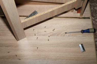 Üst görüntüden gösterilen parçacık tahtasından yapılmış mobilya montajında hafif ahşap paneller, dağınık vidalar ve bir tornavida bulunuyor. Bu DIY onarımı, ev geliştirmesi veya inşaat konuları için ideal bir örnektir.