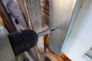 Bu, iş yerindeki kırmızı elektrikli matkabın detaylı bir yakın çekimi, eski, kırık ahşap bir duvara karşı metal bir pencere çerçevesinin kendi kendini tıklatan bir vida vidası.. 