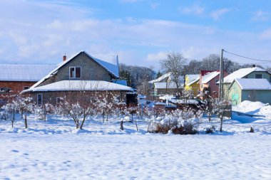 Kırsal alanda, karlı bir kış günü. Karla kaplı çatıları olan birçok ev, çıplak çalılar ve ağaçlarla çevrili, parlak mavi bir gökyüzünün altında dizilmiş. Sahne, dinginlik ve el değmemiş doğa atmosferini çağrıştırıyor..