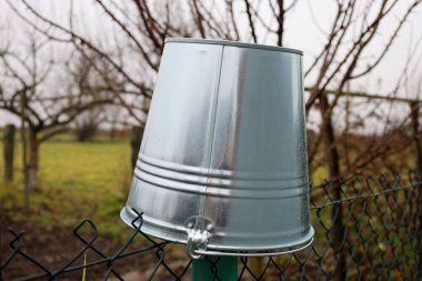 Baş aşağı duran metal galvanizli bir kova tel örgü çitlere asılı, dışarıda, uzun bir direkte dinleniyor. Gri, bulutlu çevreyi yansıtır ve bir sonbahar tarlasının arka planında yağmur damlalarını gösterir..