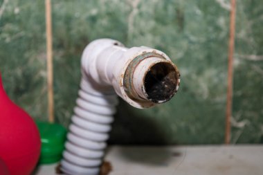 Yakın plan, çok kirli ve muhtemelen tıkanmış bir iç kanala bağlı kıvrımlı plastik bir su boşaltma borusu gösteriyor. Etrafında kir izleri görülüyor. Arka planda yeşil bir mermer fayans fark ediliyor. Tesisat sorununu işaret ediyor..