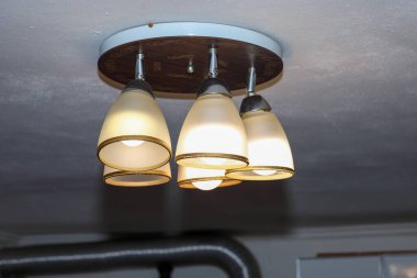 Modern bir tavan avizesi, sıcak, sarı bir ışıkla odayı hafifçe aydınlatan üç kremalı gölgeye sahiptir. Armatür ahşap bir zemine bağlı, rahat bir atmosfer yaratıyor ve iç detayları vurguluyor..