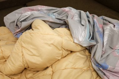 Yumuşak sarı bir yorgan ve hassas tüy desenli gri bir yatak örtüsü dağınık bir yığının içinde yatar, ev konforu ve sabah rahatlığı ortamı yaratır..