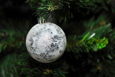 Zarif, beyaz bir Noel balosu. Yumuşak, gümüşümsü bir deseni var. Arkaplanda yeşil iğneler asılı..