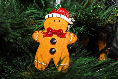 Kırmızı şapka takan ve çam ağacından bir dala asılı gülümseyen zencefilli kurabiye şeklinde parlak bir Noel süsü. Bu detaylı şenlik dekoru, kış tatili için ev konforu ve büyülü bir beklenti ortamı yaratıyor..