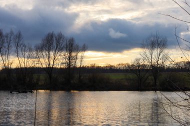 Alacakaranlıkta sakin bir gölün manzarası yoğun mavi bulutların altında altın gün batımını aydınlatan bir ışık. Kıyıdaki ince ağaç siluetleri ve su yüzeyindeki yumuşak yansımalar huzur ve doğal bir atmosfer yaratıyor.