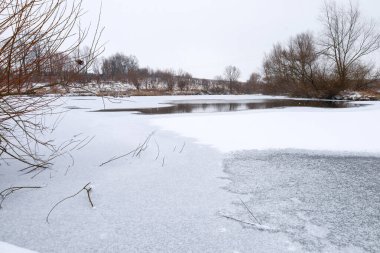 Geniş donmuş bir gölet bulutlu bir gökyüzünün altında uzanır, sakin bir kış mevsiminde çıplak ağaçlar ve karlı kıyılarla çevrilidir..