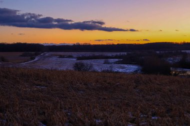 Resmedilmeye değer bir kış günbatımı kar tozlu tepeler ve tarlalar üzerinde gökyüzünü canlı turuncu ve mor renklerle boyar. Ufuktaki kara bulutlar ve orman siluetleri akşam doğasında dramatik ve barışçıl bir atmosfer yaratıyor.
