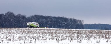 Lviv, Ukrayna - 07 Ocak 2026: Gri bir gökyüzü altında yoğun bir kış ormanına karşı karlı bir tarlada güçlü bir tarım biçicisi duruyor.