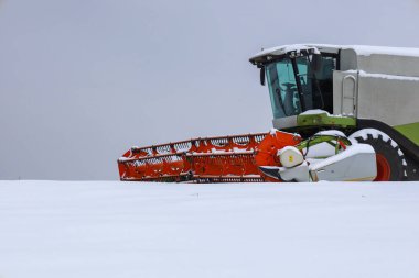 Modern bir tarım, parlak kırmızı bir başlıkla birleşir. Gri bir kış gökyüzünün altında, karla kaplı bir tarlada durur..