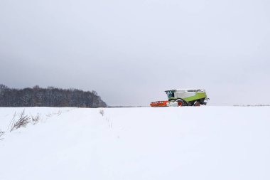 Modern bir tahıl hasatçısı sert kış ayazları sırasında karlı bir yamaçta karmaşık tarla çalışmaları yapar. Güçlü makineler, engin beyaz bir arazide ekin kalıntıları topluyor.,