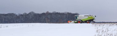 Modern yeşil bir biçerdöver, kasvetli kış gökyüzünün altındaki karlı bir tepede hasat işini tamamlar. Geniş ölçekli panorama, güçlü tarım makineleri ve geniş beyaz alan arasındaki zıtlığı vurguluyor.,