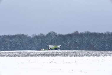 Güçlü bir yeşil biçerdöver, beklenmedik kış ayazları sırasında karla kaplı bir arazide çalışır. Tarım makineleri, buzla kaplı bir ormana karşı beyaz bir arazide çalışır.,