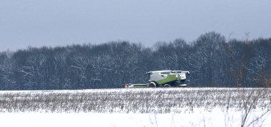 Modern bir tahıl hasatçısı, gri bir kış göğü altında yoğun karla kaplı bir ormana karşı tarla çalışması yapar. Bu geniş açı, endüstriyel tarım makineleri ile geç hasat sırasında donmuş kış doğası arasındaki zıtlığı gösteriyor..
