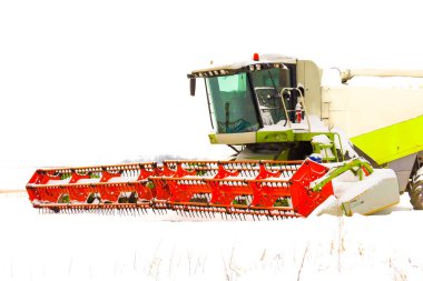 Büyük turuncu başlıklı modern bir tahıl biçme makinesi, karla kaplı bir alanda yeni tarım sezonunu bekliyor. Güçlü tarım makineleri kısmen taze karla kaplıdır, soğuk kış ile parlak bir kontrast yaratır.