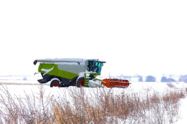Mevsim bittikten sonra modern bir tahıl biçicisi karla kaplı bir tarlanın ortasında duruyor. Parlak tarım makineleri beyaz kar ve ön plandaki kuru otlarla çelişiyor..