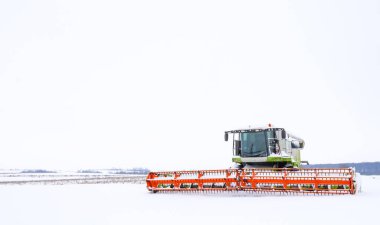 Karlı bir ovanın ortasında, bulutlu bir kış gökyüzünün altında büyük bir tahıl hasatçısı yeni mevsimi bekliyor. Parlak turuncu ekipmanlar minimalist beyaz manzaraya karşı durarak güçlü bir görsel aksan yaratıyor..