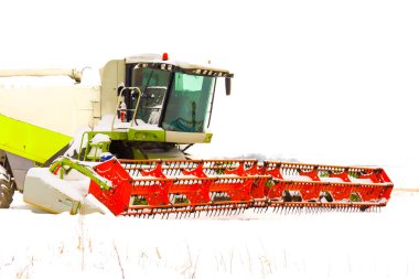 Açık bir arazide kış mevsiminde modern bir tahıl hasatçısı. Parlak turuncu başlıklı güçlü tarım makinesi kısmen taze karla kaplı, yaklaşan hasat mevsimi için teknolojik dayanıklılığı ve hazır olmayı simgeliyor.