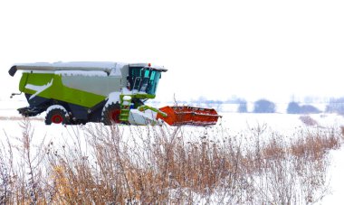 Karlı bir tarlanın kenarında kış mevsiminde depolanan modern bir tahıl hasatçısı. Ön planda, kuru otlar ve yabani otlar karları dürterek güçlü tarım makineleri için doğal bir çerçeve oluşturuyor..