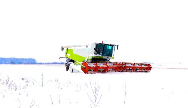 Kış uykusunda geniş bir kar tarlasının ortasında yakalanan modern bir tarım biçicisinin geniş bir görüntüsü. Makinenin beyaz-yeşil gövdesi ve parlak turuncu başlık oluşturur