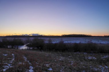 Karlı bir tarlanın resimli panoramik görüntüsü ve alacakaranlıkta, yumuşak bir eğimin altında uzak bir orman çizgisi. Huzurlu akşam atmosferi, kış bölgelerinin doğal güzelliğini vurgular.,
