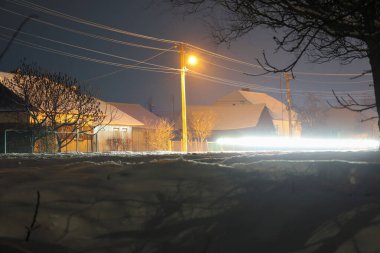 Kar altında evleri ve karanlık bir gökyüzüne karşı parlak sokak feneri ışığı olan samimi bir köyün kış gecesi manzarası. Uzun pozlama, kar ve çıplak ağaçların yanından geçen araba farlarından dinamik bir iz yakalar..