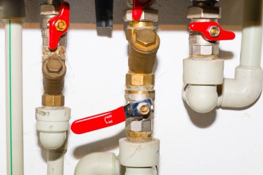Pirinç bağlantıları ve kırmızı kapama vanaları olan karmaşık bir su boruları sistemi beyaz bir duvara dayalı. Tesisat ünitelerinin detaylı görünümü, profesyonel tesisat kurulumunu gösterir,